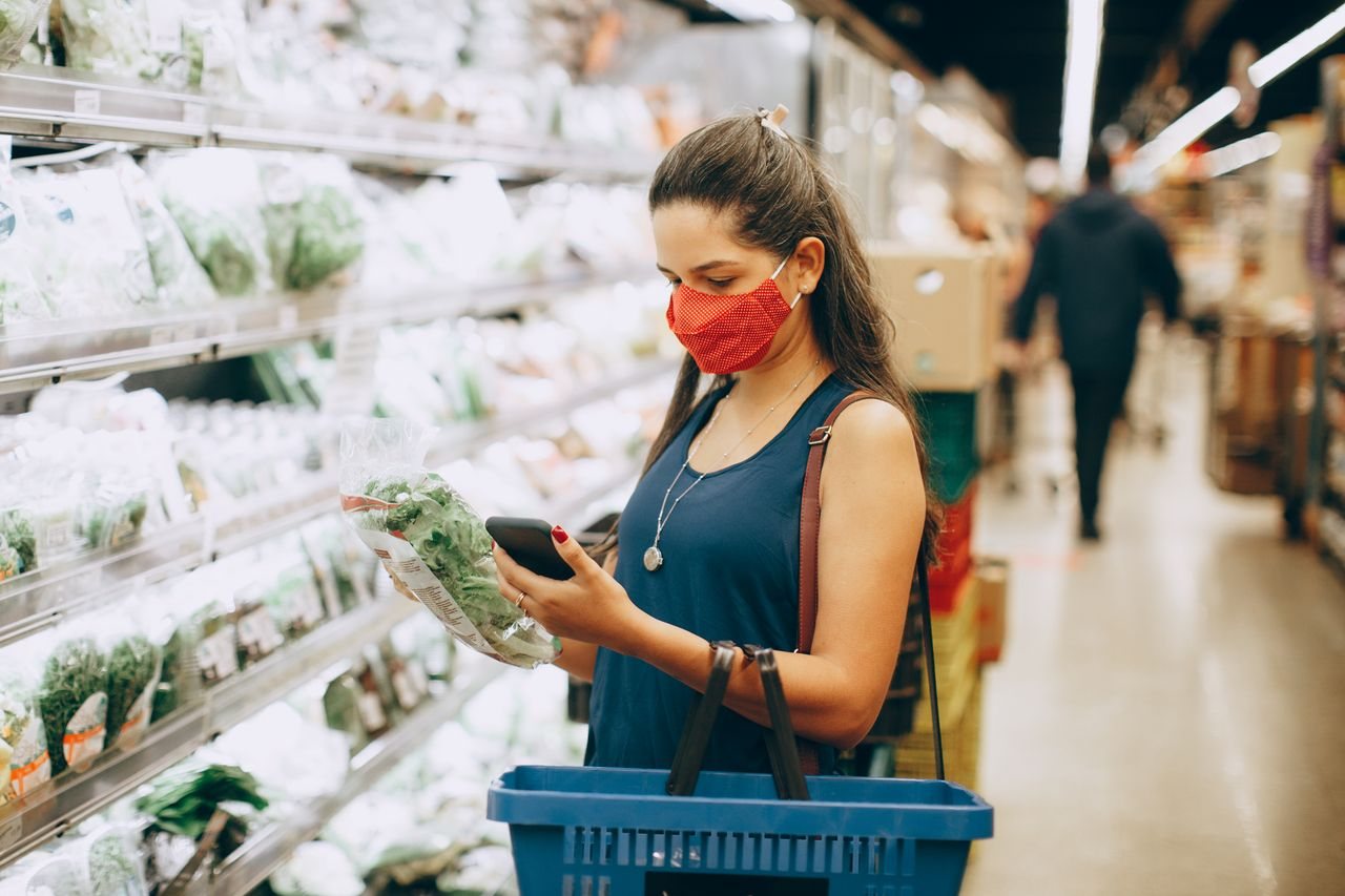 Free A woman wearing a mask shops for groceries in a supermarket, checking products with her phone. Stock Photo